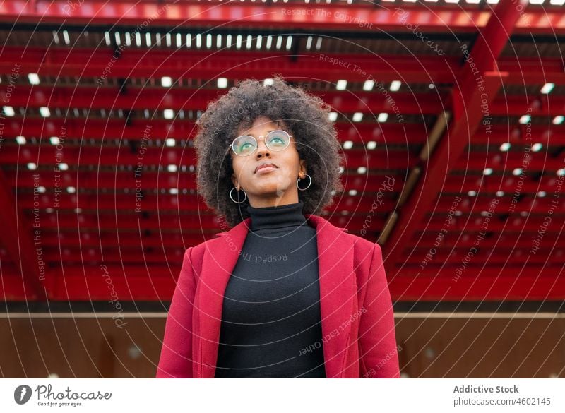Afroamerikanische Frau in karmesinrotem Mantel und mit Brille Afro-Look Frisur positiv krause Haare lässig Porträt charmant Optimist Menschliches Gesicht