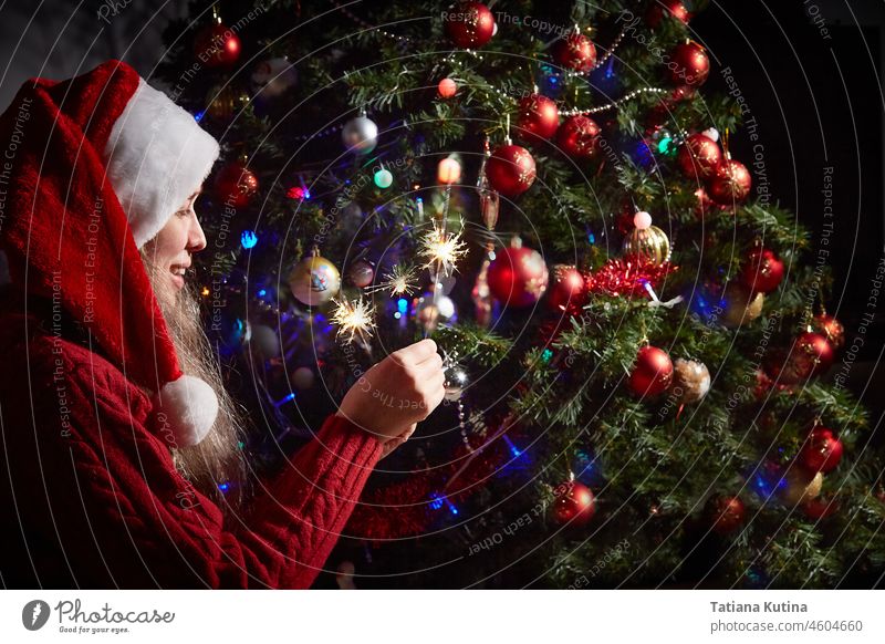 Frau mit Weihnachtsmannmütze und Wunderkerzen vor dem Hintergrund eines mit roten Kugeln geschmückten Neujahrs- oder Weihnachtsbaums. feiern Weihnachten Party