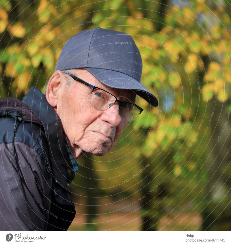 Porträt eines Senioren mit Kappe in der Natur, der etwas skeptisch zur Seite blickt Mensch Mann Blick Brille Jacke draußen Baum Herbst Herbstfärbung