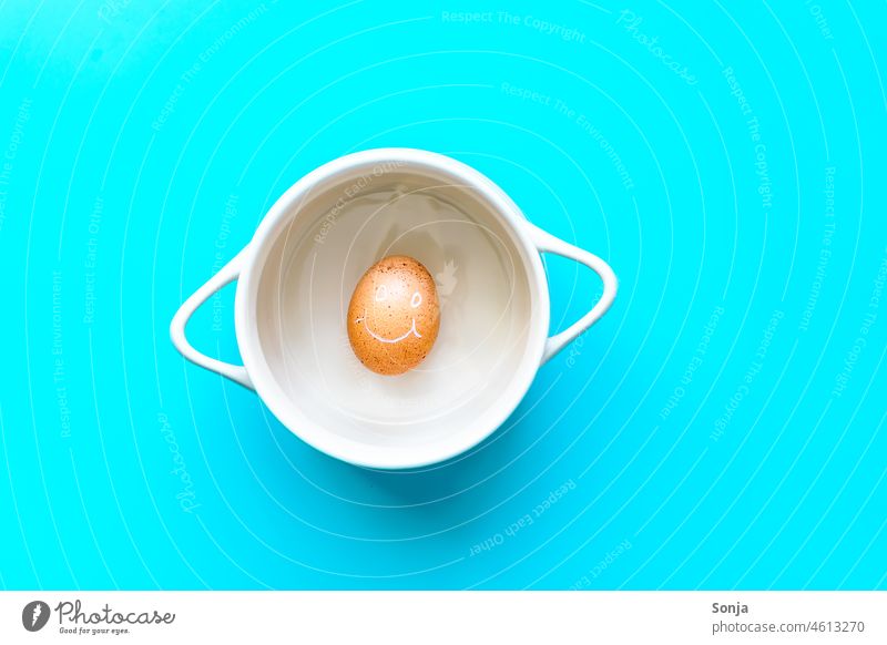 Ein Ei mit lächelnden Gesicht in einem Kochtopf auf einem blauen Hintergrund. Draufsicht. Hühnerei braun Lächeln Frühstück Protein Humor Bioprodukte Eierschale