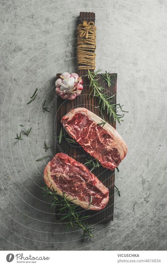 Zwei roh marmorierte Rib-Eye-Steaks auf Schneidebrett mit Rosmarin, Knoblauch und grünem Pfeffer Rippenauge zwei marmoriertes Steak Paprika Beton Küche Tisch