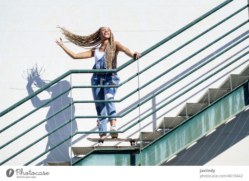 Schwarzes Mädchen auf einer städtischen Treppe, das ihre bunten Zöpfe im Wind bewegt. Typisch afrikanische Frisur. Frau schwarz Zopf afrobraid Rastalocken