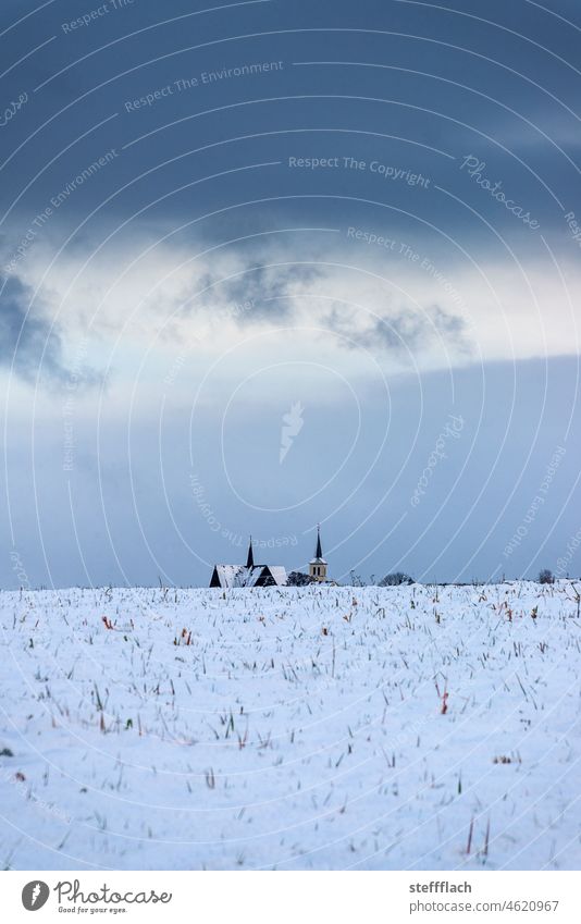 Zwei Kirchen hinter schneebedeckten Feldern vor dunklem wolkigen Winterhimmel Wolken dunkel düster kalt Himmel Blau Dunkelblau Grau Dämmerung Abend