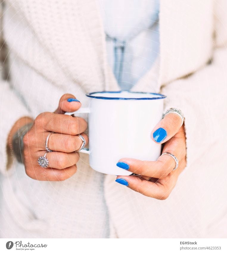 Nahaufnahme der Hände einer jungen Frau mit blau lackierten Nägeln und Fingerringen, die einen weiß emaillierten Becher mit Kaffee oder Tee hält lackierte Nägel