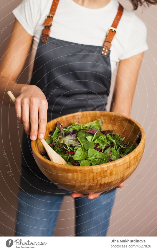 Frau in einer grauen Schürze mischt grünen Salat in Öko-Holz-Bambus-Schüssel in der Küche. Gesundes Essen und Ernährung, umweltbewusster Lebensstil.
