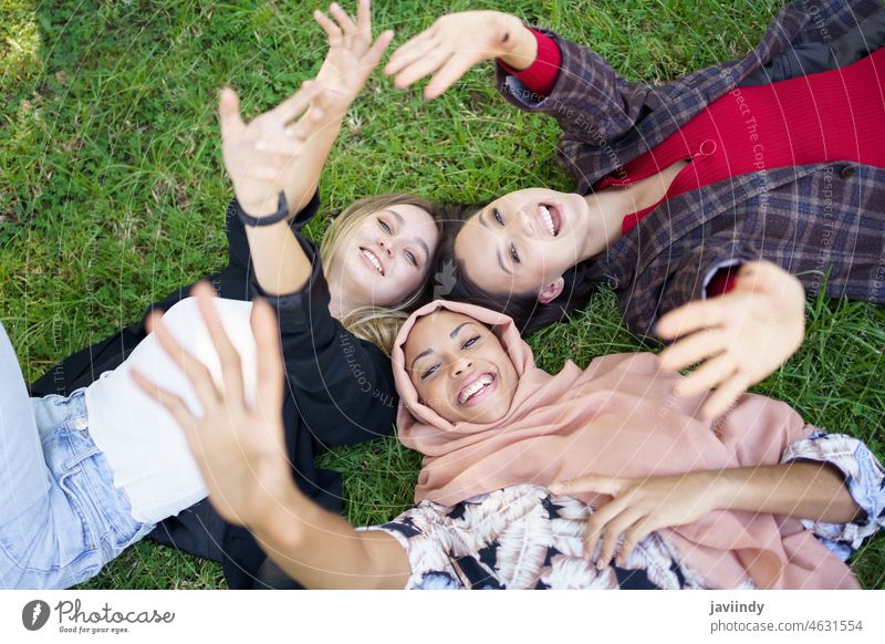 Fröhliche junge diverse weibliche Freunde liegen auf Gras und lächeln in die Kamera Frauen Lächeln Lügen Rasen Park Zusammensein Wellenhand Freundschaft