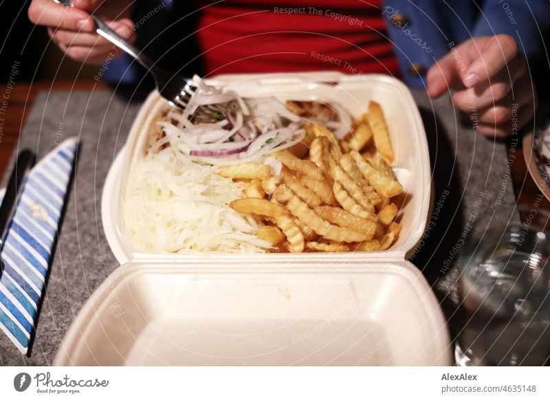 Frau mit rotem Pullover und blauer Jacke sortiert Zwiebelstreifen aus einer Styropor- Menübox aus einem Gyrosteller mit Pommes und Krautsalat - Abendessen vom Lieferservice - Die kleine Kollossos- Platte