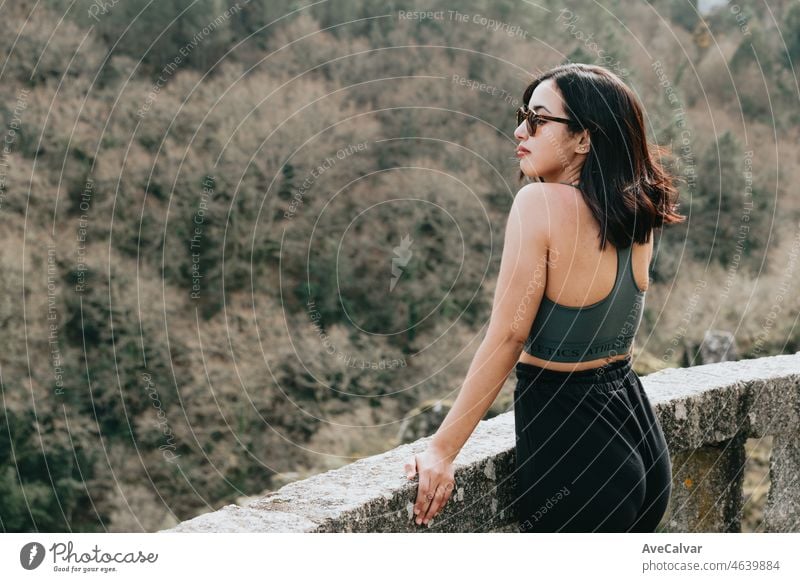 Eine junge Frau Reisende in sportlicher Kleidung und Sonnenbrille mit Blick von einer Klippe in den Wald, bewundern die Aussicht auf die Tierwelt. Gesundes Leben in der Natur junge Menschen Konzept.