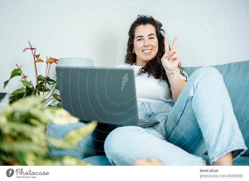 Junge Freiberufler latina Frau arbeitet am Computer, während auf dem bequemen Sofa zu Hause sitzen lächelnd in die Kamera tun Sieg Zeichen auf moderne trendige Kleidung. Konzept der Remote-Arbeit von zu Hause aus