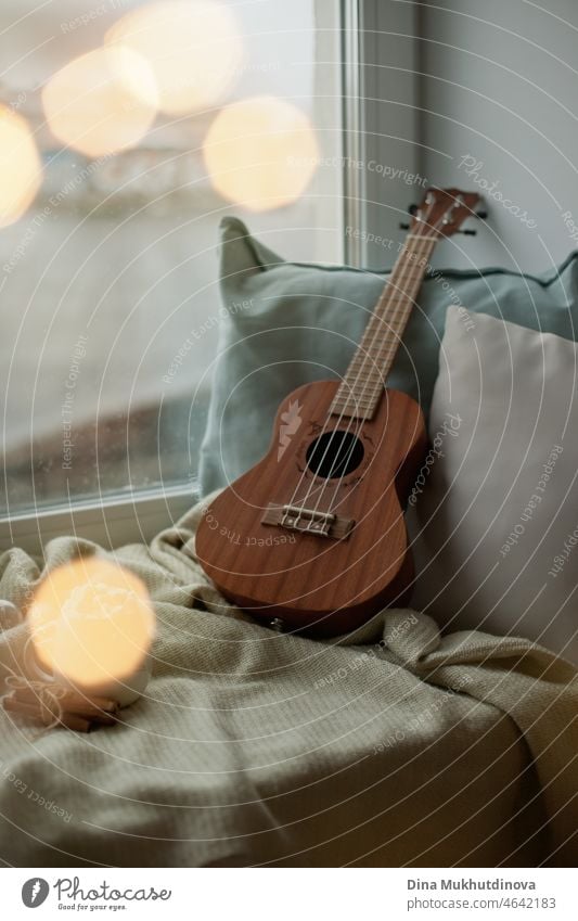 Ukulele zu Hause auf der Fensterbank mit kuscheligen Decken und Kissen an einem kalten Regentag. Gemütliche Stimmung, die Zeit zu Hause verbringen. Melodie und Musik Hobby - kleine Gitarre zum Musizieren.