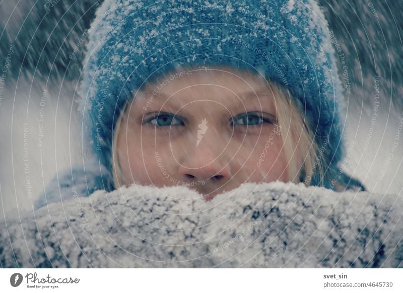Porträt eines blonden Mädchens mit blauer Strickmütze und verschneiten Fäustlingen bei starkem Schneefall. Es schneit. im Freien Winter weiß blondes Mädchen