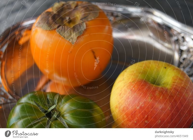 Stillleben aus Obst und Gemüse in Form einer Persimon oder einer Khaki, eines Apfels und einer grünen Tomate auf einem silbernen Tablett Ernährung Einsamkeit