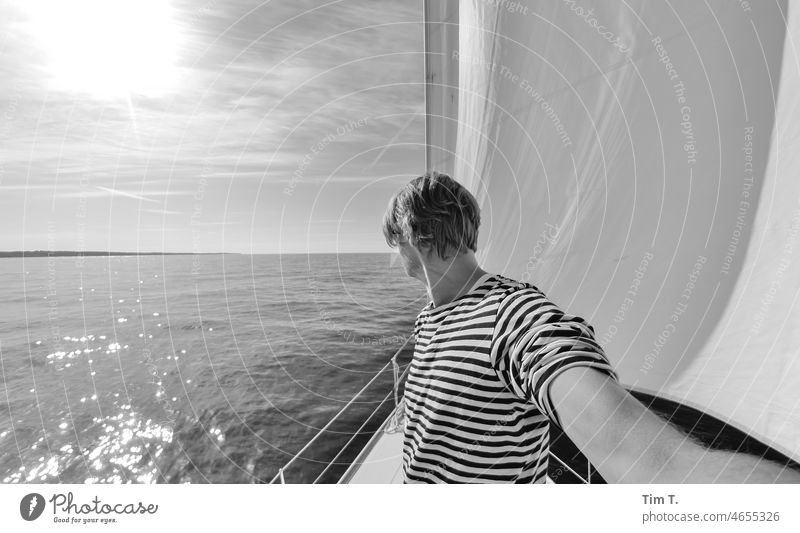 ein Mann steht am Bug eines Segelschiffes Segeln s/w Schwarzweißfoto Tag Außenaufnahme b/w b&w Ostsee Segelboot gestreiftes hemd Sonnenlicht kein wind ruhig