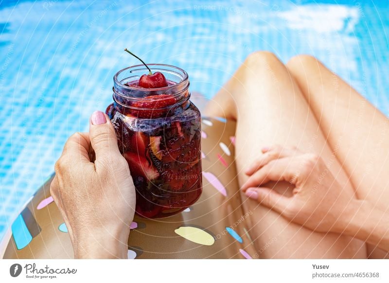 Schöne Frauen in einem blauen Swimmingpool. Die anmutige Hand einer Frau hält einen Beerencocktail. Aufblasbarer Donut. Nahaufnahme. Sommerurlaub Stockfoto. Rückansicht.