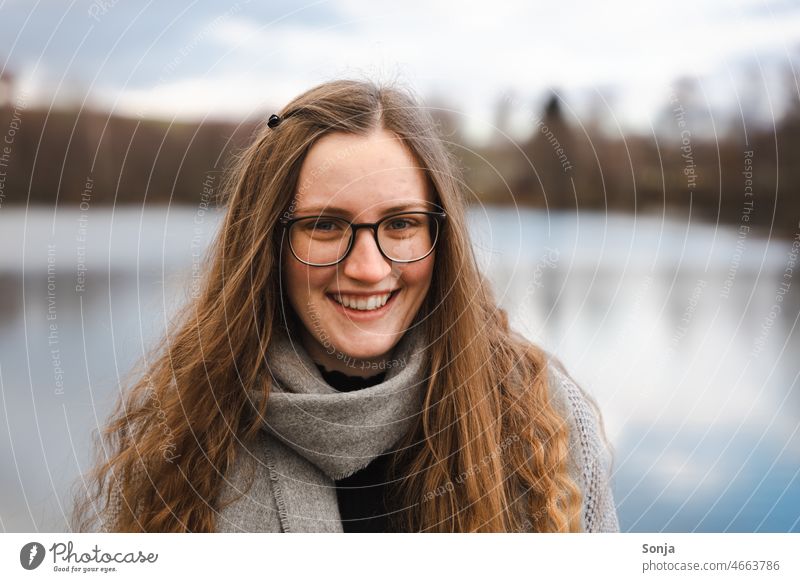 Eine junge lächelnde Frau mit Brille und langen Haaren an einem See. Junge Frau Lächeln feminin Porträt lange Haare hübsch attraktiv Außenaufnahme Glück