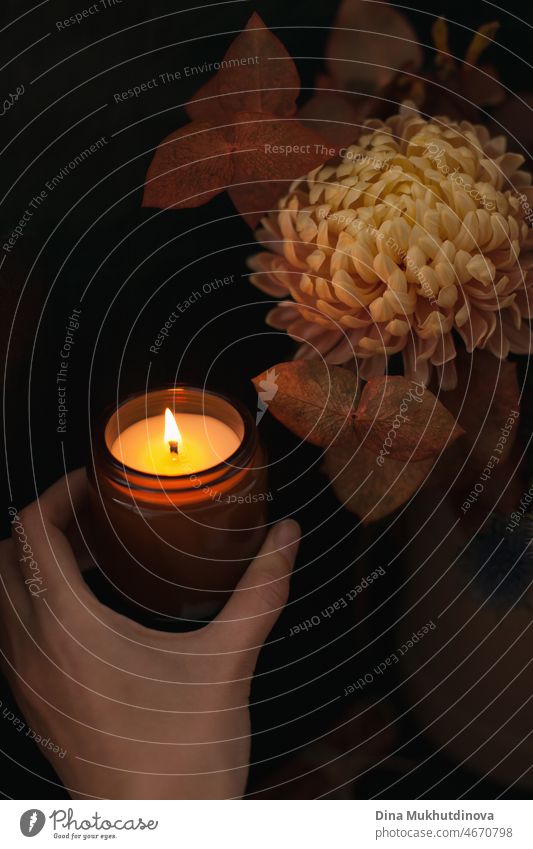 Hand hält eine Duftkerze in einem braunen Glasgefäß in der Nähe von Blumenstrauß, Aromatherapie und Spa. Glühende brennende Kerze und Blumen braunen Hintergrund. Spa, Aromatherapie und behandeln Sie sich selbst Konzept.