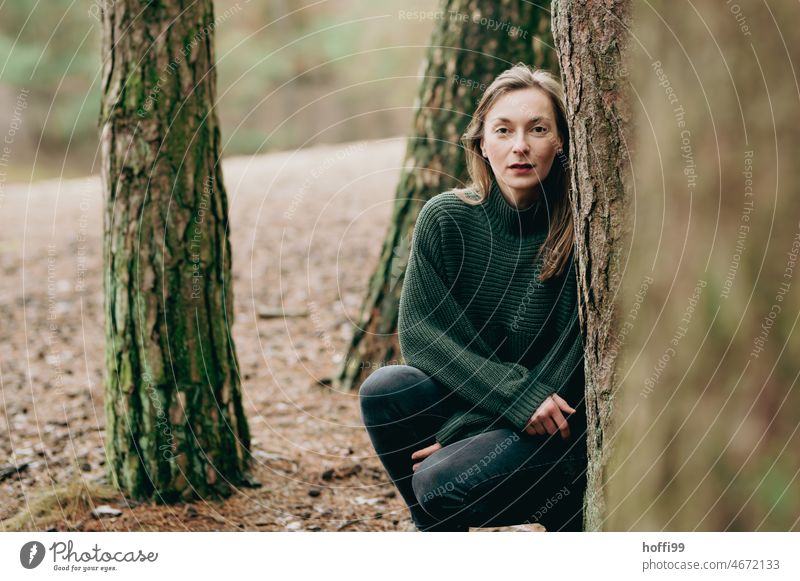 eine Frau hinter Bäumen blickt gespannt in die  Kamera Blick Junge Frau melancholisch Porträt besinnlich ruhig nachdenklich Wald Haarsträhne Natur Frauengesicht