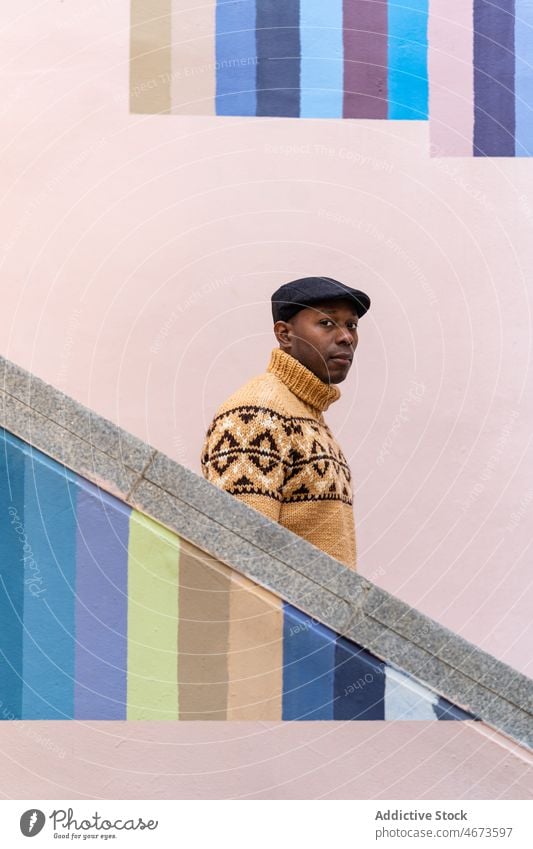 Schwarzer Mann in warmem Pullover geht die Treppe hinunter warme Kleidung im unteren Stockwerk trendy Verschlussdeckel gestrickt farbenfroh Streifen Wand lässig
