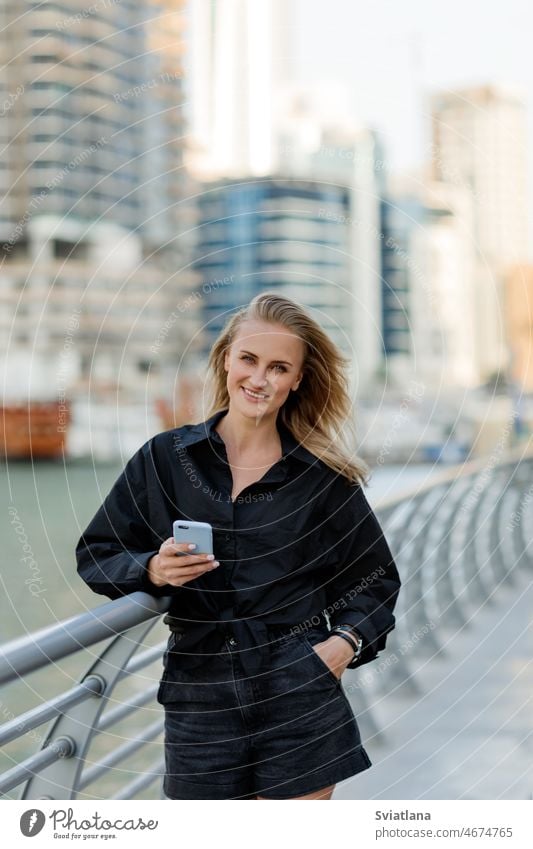Eine attraktive Frau steht am Ufer und benutzt ihr Telefon, während sie durch die Stadt spaziert blond Straße Stauanlage stylisch schön Urlaub Lächeln benutzend