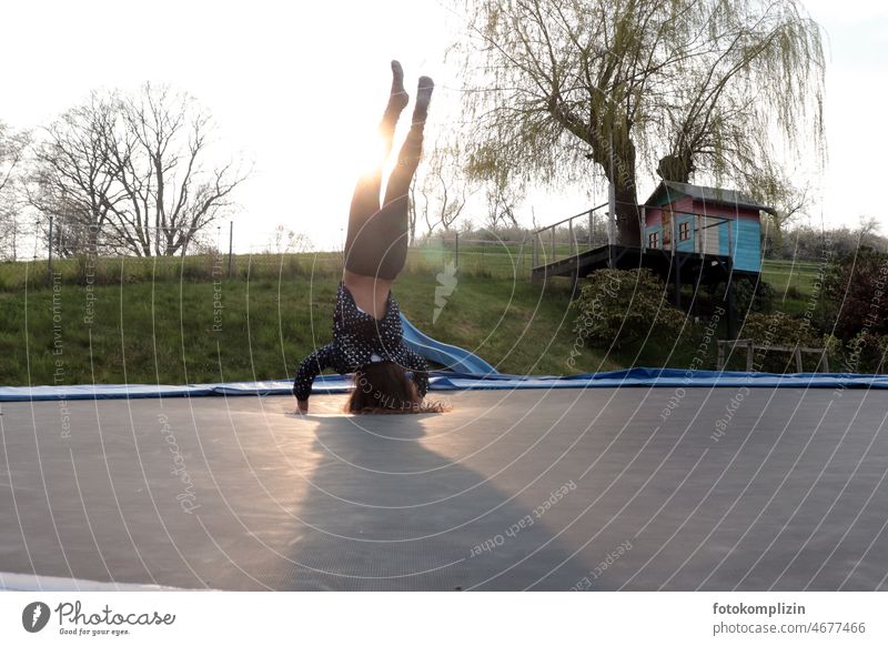 Frühlingsanfang: Mädchen macht Kopfstand auf einem Trampolin mit Baumhaus im Hintergrund Kind spielen bewegen sich freuen alleine Frühlingsgefühle Kinderspiel