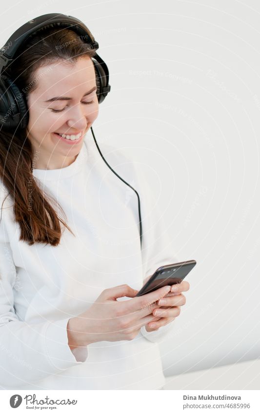 Junge brünette Millennial-Frau, die ein Mobiltelefon benutzt und mit Kopfhörern Musik hört, lächelnd.  Junger Erwachsener in weißem Sweatshirt mit Technologie isoliert auf weißem Hintergrund.