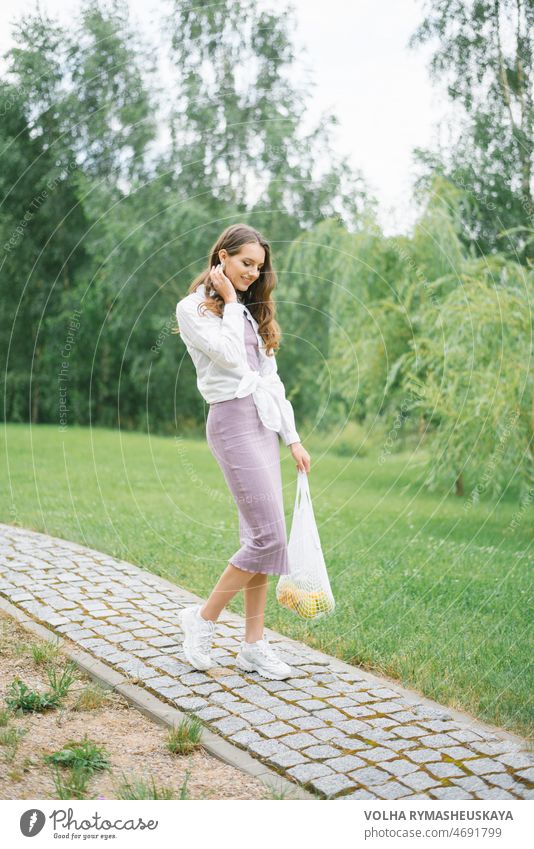 Stilvolle glückliche Frau, die einen Weg in einem Park entlanggeht und Obst in einer Netztasche trägt Model Spaziergang Spaß Shorts Weiblichkeit passen viele