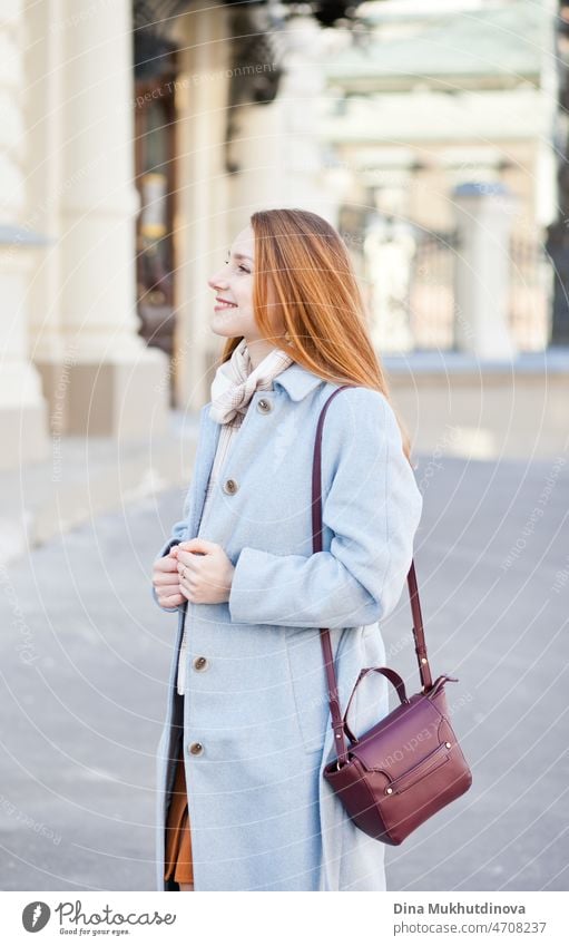 Junge schöne Frau in einem hellblauen Mantel steht auf der Straße in der Stadt im Sonnenlicht an einem sonnigen Tag. Candid Lifestyle-Porträt einer Frau, lächelnd.