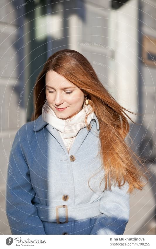 Junge schöne Frau in einem hellblauen Mantel steht auf der Straße in der Stadt im Sonnenlicht an einem sonnigen Tag. Candid Lifestyle-Porträt einer Frau, die in die Kamera schaut und lächelt.