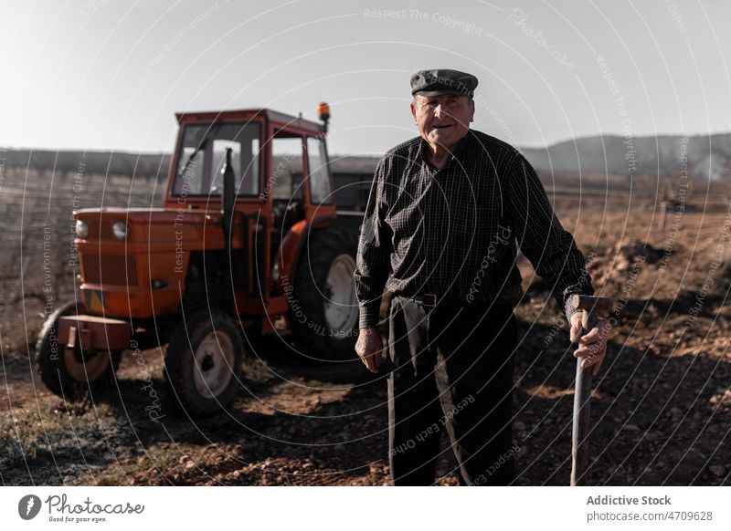 Lächelnder älterer Mann mit Gartengerät in der Nähe eines Traktors auf einem Feld Landwirt Ackerbau Karre Bauernhof ländlich Arbeit Landschaft männlich Verkehr
