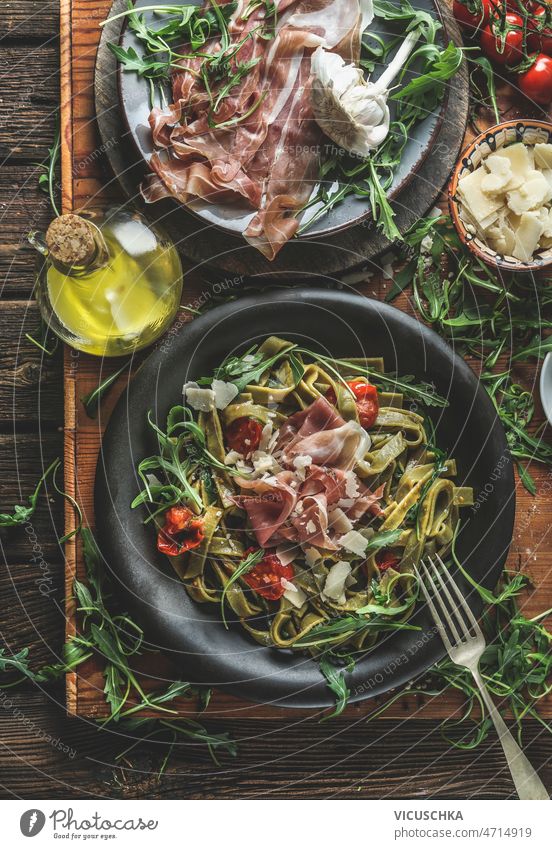Grüne Tagliatelle-Nudeln mit Schinken, Parmesan, Rucola und Tomaten auf schwarzem Teller grün Spätzle Käse rustikal hölzern Küche Tisch Zutaten Olivenöl Besteck