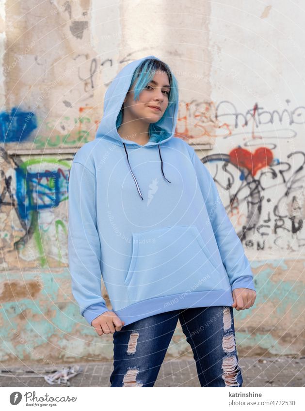 Blauhaariges Teenager-Mädchen in hellblauem Oversize-Kapuzenpulli, das an einer Graffiti-Wand steht hell-blau Attrappe Übergröße Jeanshose blauhaarig im Freien