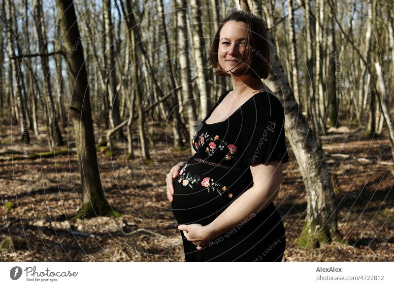 Junge, schwangere Frau steht in schwarzem Umstandskleid im Wald junge Frau schön Schönheit neues Leben Kleid barfuß Schwangerschaftsfoto Bauch Babybauch