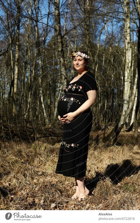 Junge, schwangere Frau steht in schwarzem Umstandskleid und Blumenkranz barfuß im Wald junge Frau schön Schönheit neues Leben Kleid Schwangerschaftsfoto Bauch