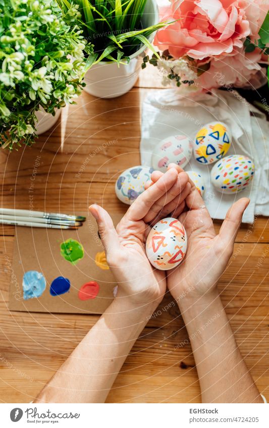 Frau hält ein modernes Osterei. Pinsel und Farben mit Blumen und Pflanzen. Glückliche Ostern Konzept auf einem hölzernen Hintergrund. Beteiligung Hand Ei Feier