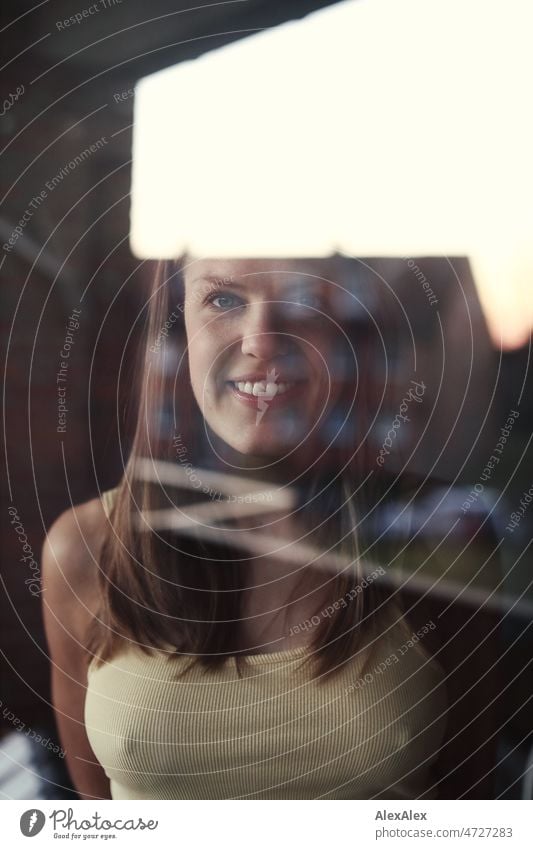 Portrait einer jungen, blonden Frau hinter einer Fensterscheibe, in der sich ein Haus und der Abendhimmel spiegeln junge Frau langhaarig lächeln Freude Gesicht