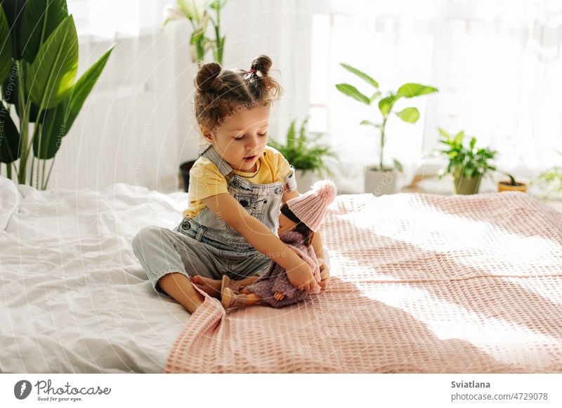 Ein kleines Mädchen sitzt auf dem Bett im Schlafzimmer und spielt mit ihrer Puppe, während sie ihr Halstuch bindet. Kind Spielzeug Kindheit Sitzen Spielen