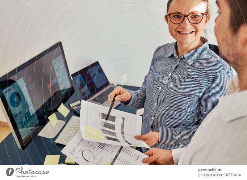 Unternehmerin bespricht Finanzdaten mit ihrem Kollegen, der am Schreibtisch im Büro steht. Glückliche, positive Geschäftsfrau, die mit Diagrammen und Tabellen am Computer arbeitet. Zwei Menschen arbeiten zusammen