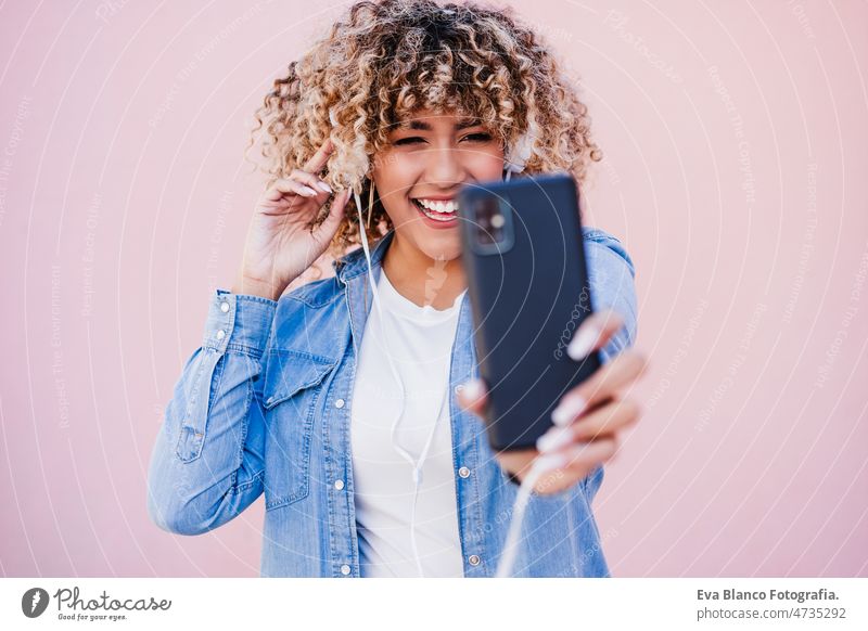 Porträt einer lächelnden hispanischen Frau mit Afro-Haar in der Stadt, die ein Mobiltelefon und ein Headset benutzt. lifestyle kurvenreich Körperfreundlichkeit