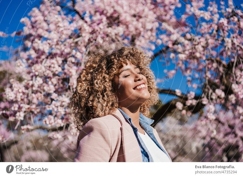 lächelnde hispanische Frau mit geschlossenen Augen im Park genießt sonnigen Tag. Frühling Blumen Hintergrund Tanzen kurvenreich Körperfreundlichkeit Afro-Look
