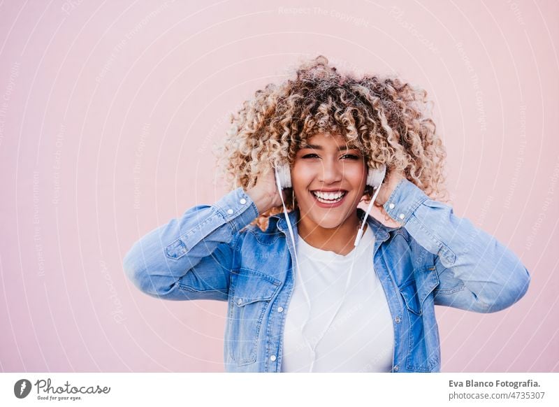 Porträt einer lächelnden hispanischen Frau mit Afro-Haar in der Stadt, die ein Mobiltelefon und ein Headset benutzt. lifestyle hören Körperfreundlichkeit