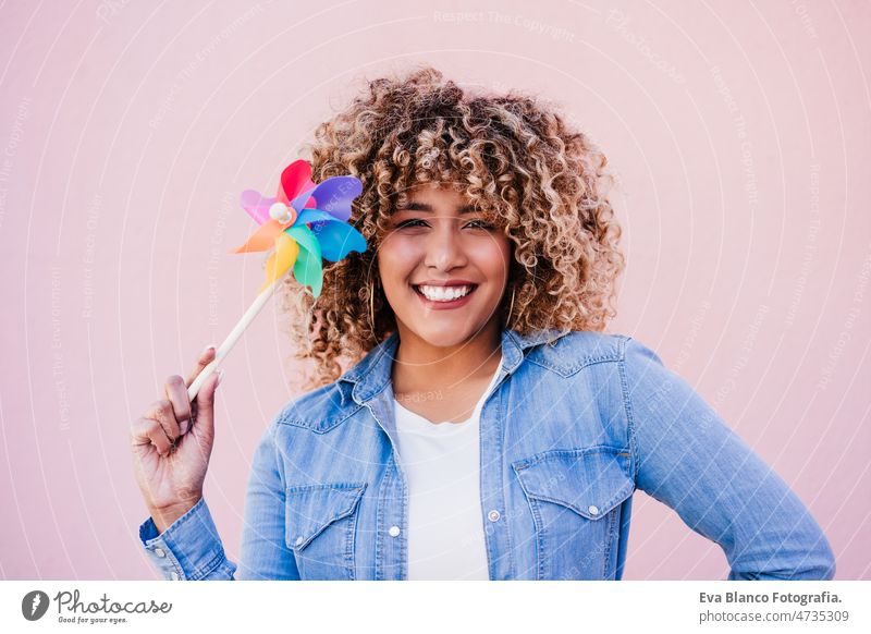 schöne glückliche hispanische Frau mit Afro-Haar hält bunte Windrad. rosa Hintergrund, Windenergie Windmühle Afro-Look farbenfroh Energie nachhaltig reduzieren