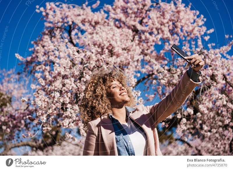 Lächelnde hispanische Frau im Park, die ein Foto mit dem Handy macht. Frühling Blumen Hintergrund Bild Technik & Technologie Smartphone Internet Gerät Tanzen