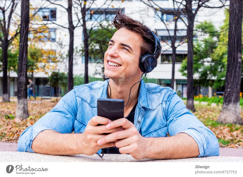 Junger Mann genießt Musik mit Kopfhörern in einem öffentlichen Park Person Mobile Sitzen Bank jung Lifestyle männlich Telefon Lächeln Glück Tagträume hören