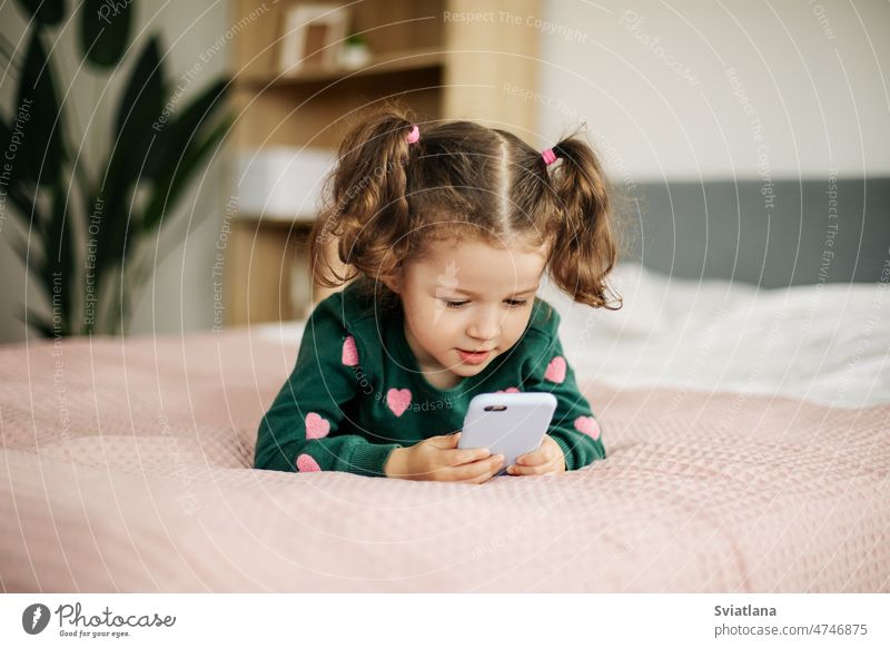 Ein kleines Mädchen schaut fröhlich auf sein Smartphone, spielt ein Spiel oder sieht sich einen Zeichentrickfilm an. Kind wenig Telefon klug Aussehen