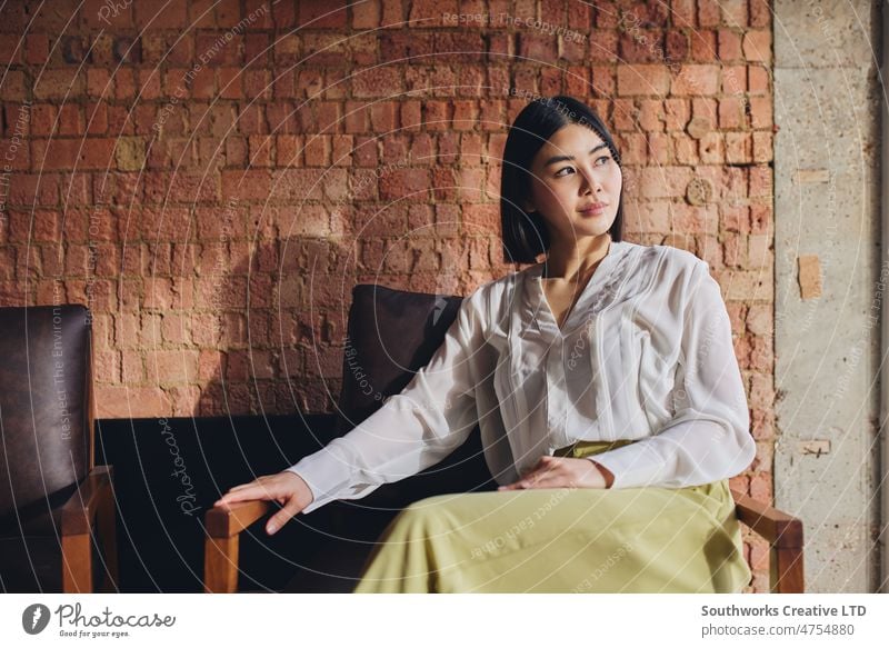 Porträt einer erwachsenen chinesischen Geschäftsfrau, die auf einem Stuhl vor einer freigelegten Backsteinmauer sitzt und wegschaut Mitte Erwachsener Chinesisch