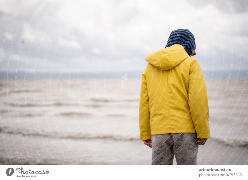 Kind in Regenkleidung am Strand bei bewölktem Wetter kind kindheit regen himmel stimmung wellen wasser wind windig windiges Wetter Wasseroberfläche Natur Wolken