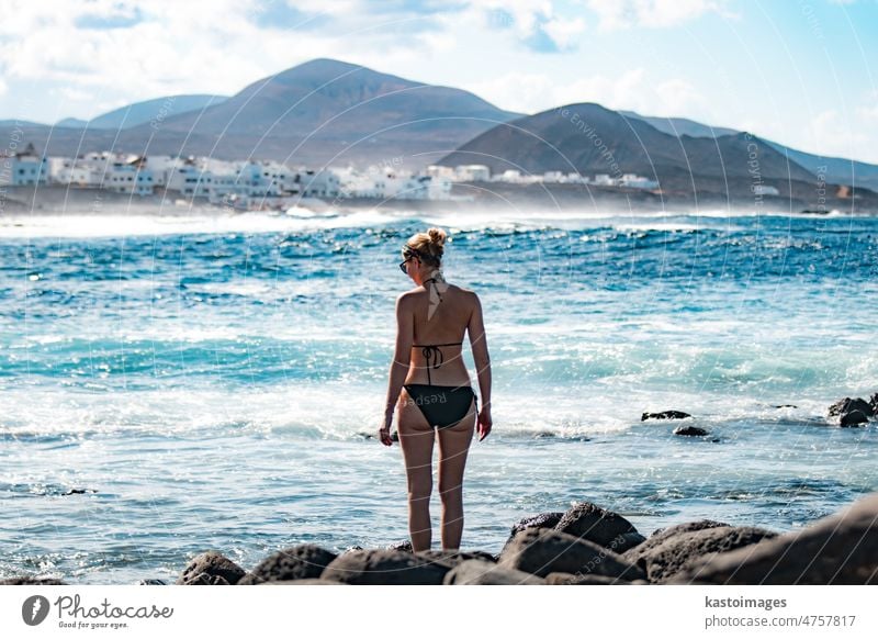 Weibliche Touristin am wilden Felsenstrand und an der Küste des Surfspots La Santa auf Lanzarote, Kanarische Inseln, Spanien. La Santa Dorf und Vulkan Berg im Hintergrund