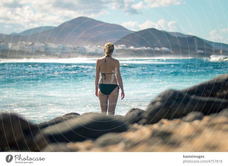Weibliche Touristin am wilden Felsenstrand und an der Küste des Surfspots La Santa auf Lanzarote, Kanarische Inseln, Spanien. La Santa Dorf und Vulkan Berg im Hintergrund