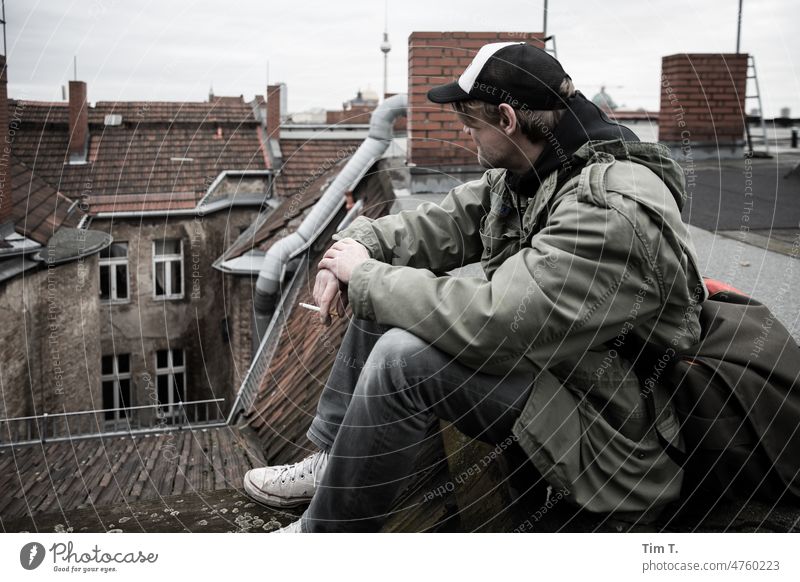 ein Mann mit Basecap und Zigarette sitzt auf einem Dach. Im Hintergrund ist der Fernsehturm zu sehen. Berlin Berlin-Mitte Berliner Fernsehturm basecap Hinterhof