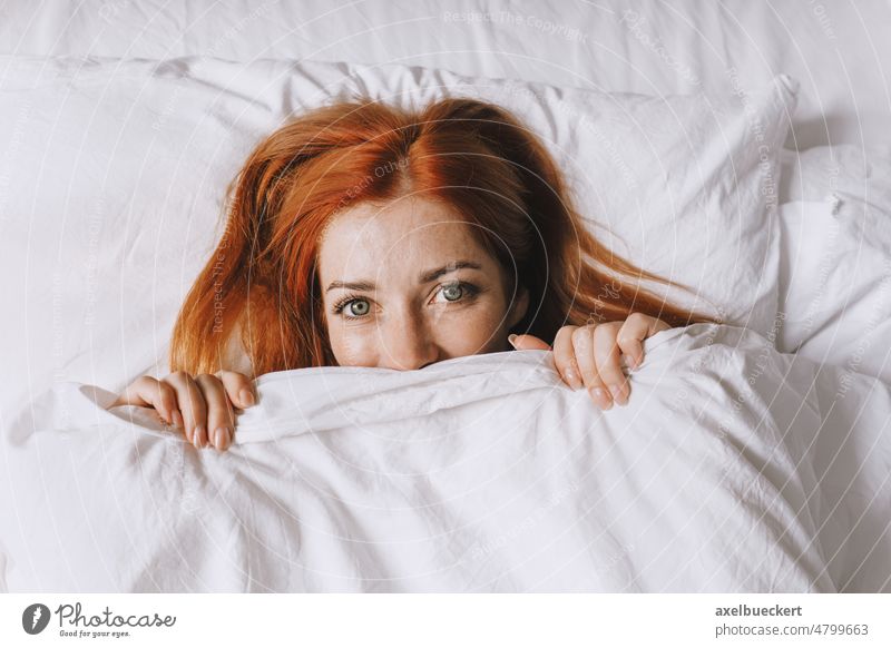 schüchterne junge Frau, die sich unter der Bettdecke versteckt zurückhaltend Mädchen Deckung versteckend unten Decke spielerisch Glück Lächeln Schlafzimmer